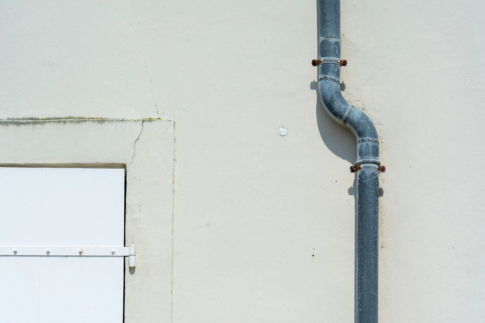 Descente d'eau pluviale fixée sur un mur enduit clair, près d'un volet